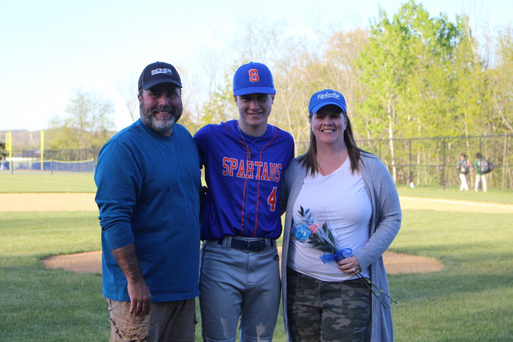 A high school boy stands between a woman on right and a man on left. He has his arms around them. He is wearing a baseball uniform, with a blue and orange shirt and gray pants. The woman is holding a flower.