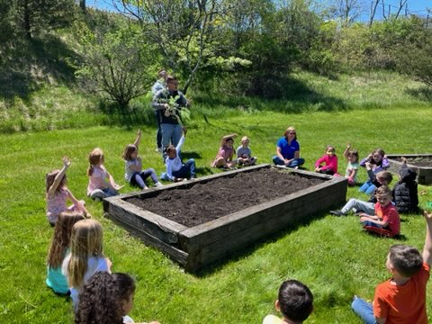 A group of about 20 kindergarten students sit around a large raised garden bed on a beautiful sunny day. There is a man at the center talking to them.