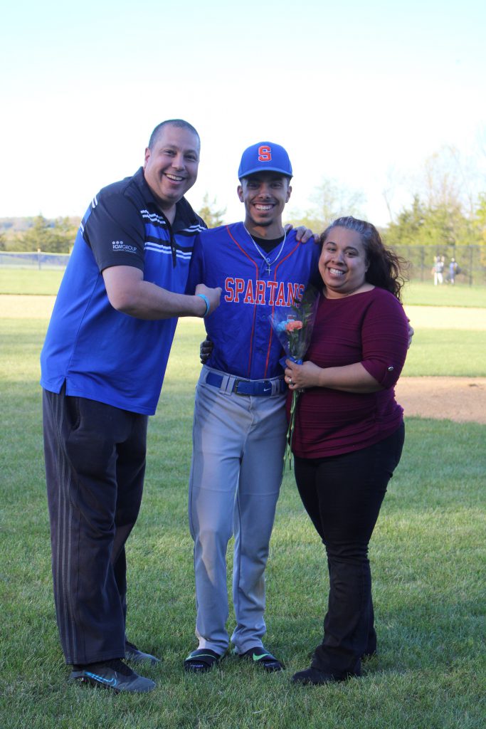 A high school boy stands between a woman on right and a man on left. He has his arms around them. He is wearing a baseball uniform, with a blue and orange shirt and gray pants. The woman is holding a flower.