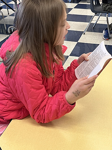 A girl with shoulder-length brown hair wearing an orange jacket has her elbows on the table and holds up her new dictionary, reading it.