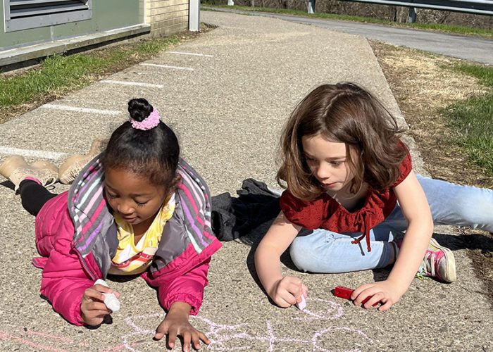 Two smaller elementary students recline on the pavement on their bellies and use chunky chalk to do math problems. The girl on the left has her dark hair up in a bun and is wearing a pink jacket. The girl on the right has shoulder-length brown hair and is wearing a red shirt and jeans.