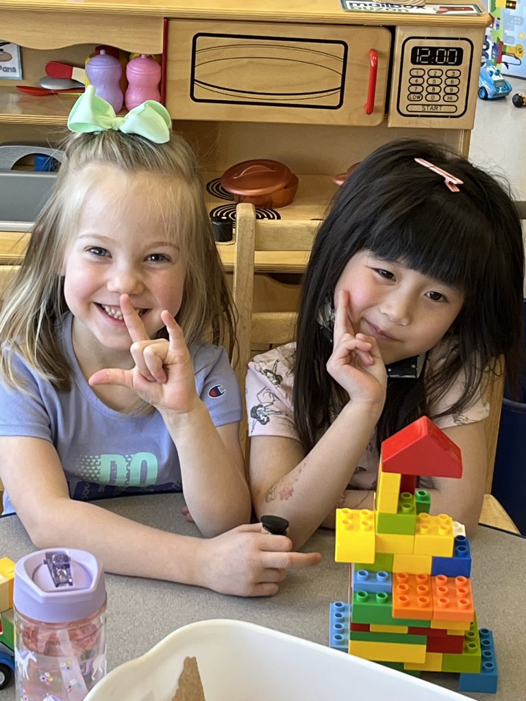 Two pre-K girls sitting at a table building with blocks. The girl on the left has shoulder-length blonde hair with a green bow and the girl on the right has long dark hair. They are both smiling.