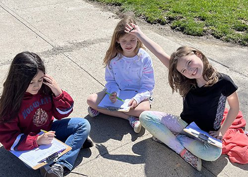 Three girls sit on the sidewalk, each with clipboards on their laps. They are holding pencils. The girl on the right is smiling and has her hand up in the air.