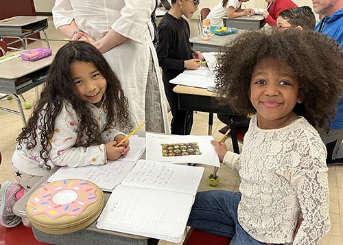 Two elementary age girls sit at a desk together with notebooks open and pencils in their hands. The girl on the left has long dark hair and is smiling. The girl on the right has shoulder-length dark curly hair. She is wearing an off-white long-sleeve shirt. Both are smiling.