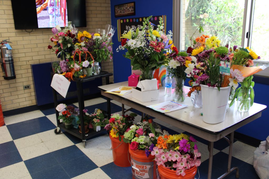 A cart with two levels that are filled with flowers. NExt to it is a table with flowers on top of it and below the table are three buckets filled with flowers.