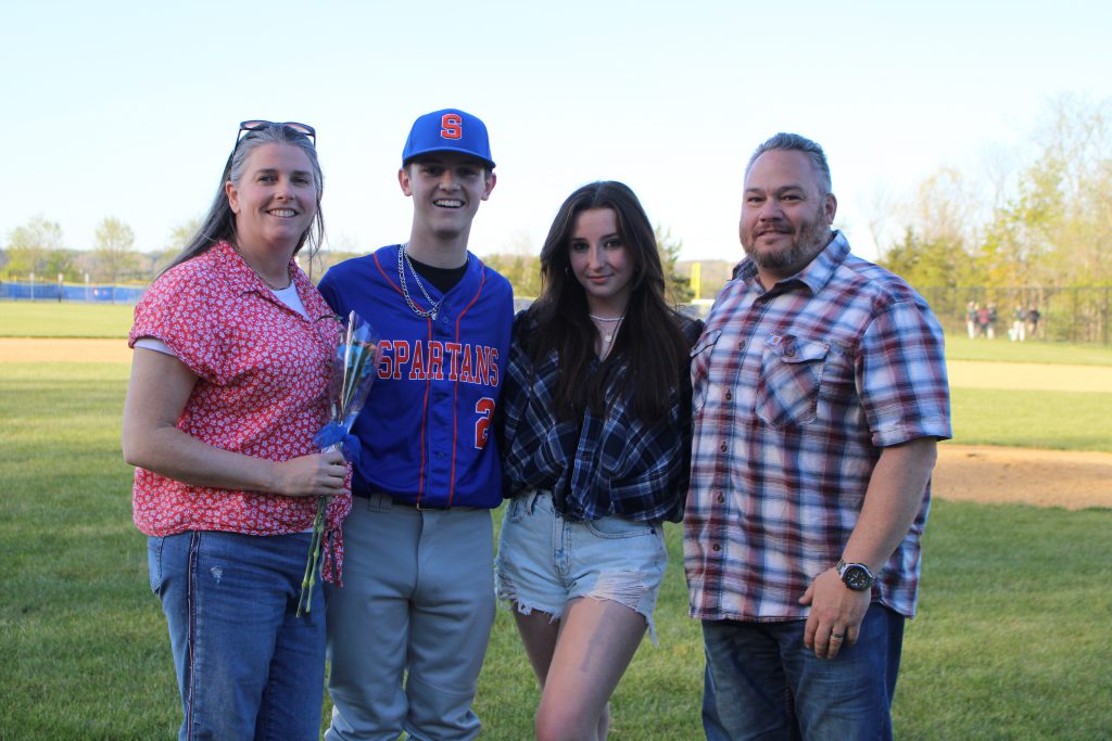 A high school boy stands with three other people, a woman onleft holding a flower, a young woman next to him, and a man on the right. He is wearing a baseball uniform, with a blue and orange shirt and gray pants.