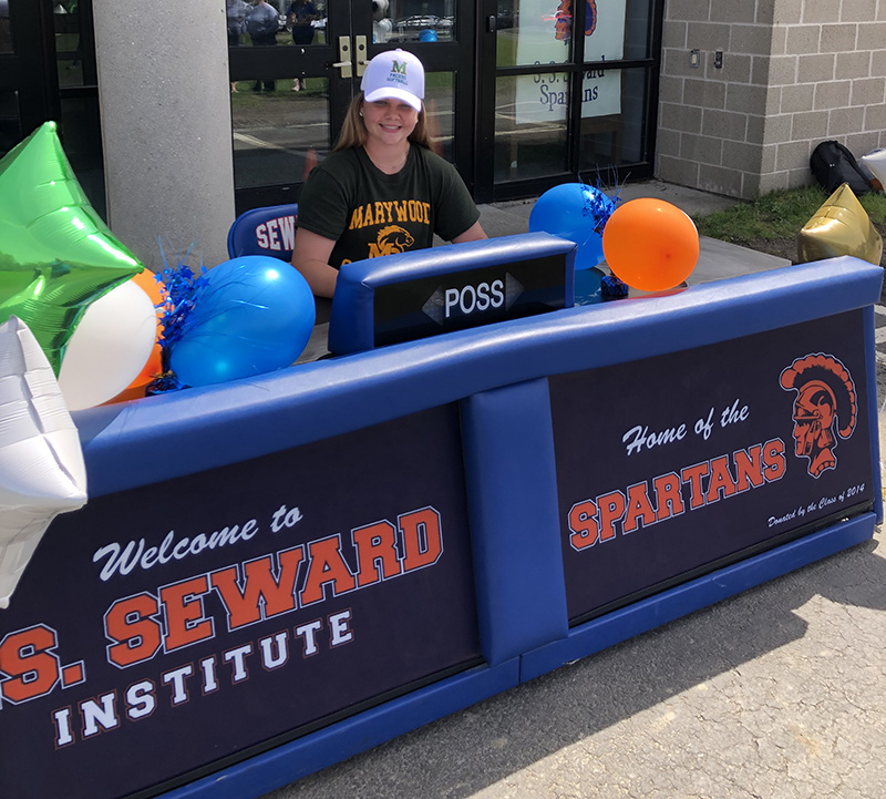 A young woman sits at a long table with blue and orange balloons and a banner in front that says SS Seward Institute. She is waring a blue tshirt that sayd Marywood and a white cap.