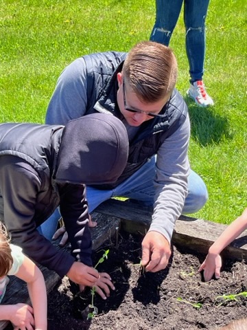 A man leans over and helps a young student plant a plant in a garden bed.