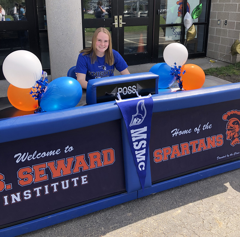 A young woman with long blonde hair wearing a blue shirt sits at a long table that has many blue, orange and white balloons. The front says S.S. Seward Institute and Home of the Spartans. 