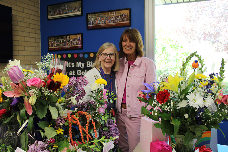 Two women stand in the center of bunches of flowers. Both are smiling.