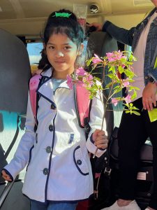 A girl wearing a button down light purple spring coat holds a spray of pink flowers. She has dark hair up in a clip and is smiling as she comes from the school bus.