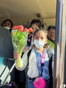 A little girl, with dark hair pulled back and a blue mask, holds a bouquet of red tulips as she comes off a school bus.