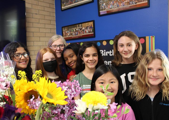 A group of seven fifth-grade girls stand with a woman.All are smiling. There are lots of flowers in front of them.