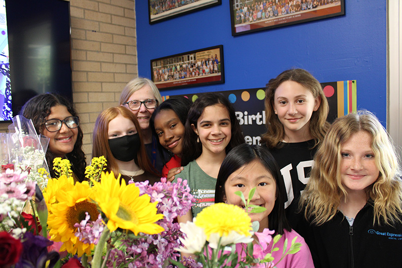 A group of seven fifth-grade girls stand with a woman.All are smiling. There are lots of flowers in front of them.