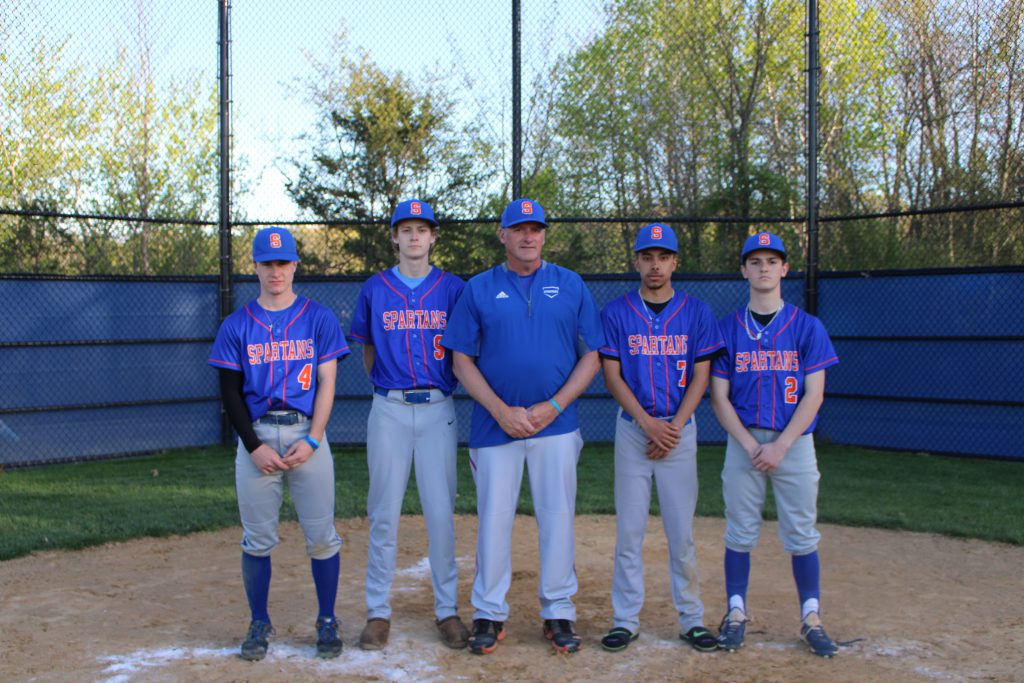 A man stands in the center of four high school baseball players. They are all wearing baseball uniforms, blue top with orange lettering that says SPARTANS,. All have blue baseball caps on.
