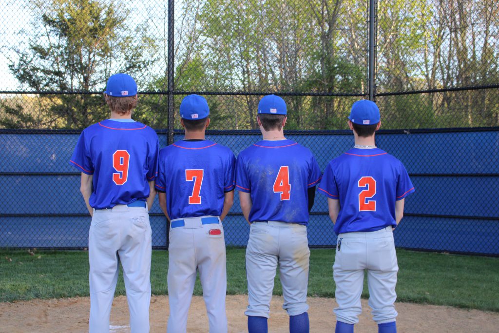 Four high school boys wearing baseball uniforms have their backs to the camera. They are wearing royal blue shirts with orange numberson them, and gray pants. they are number 9, 7, 4 and 2 from left.