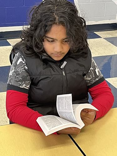 A fourth-grade student with shoulder-length dark hair, wearing a red long-sleeve shirt and black vest, pages through a paperback dictionary.