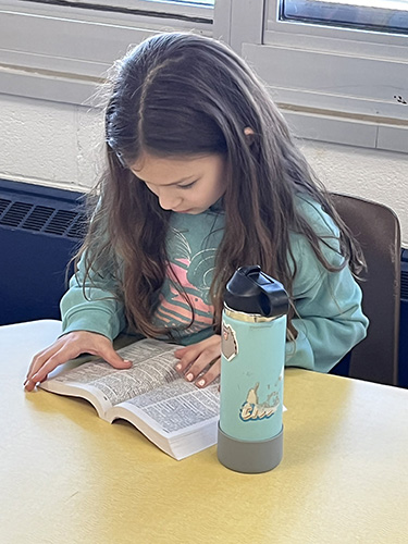 A girl with long dark hair, wearing a green sweatshirt has a paperback dictionary open on her desk. He is pointing to words in the dictionary. There is a green water bottle on the desk as well.