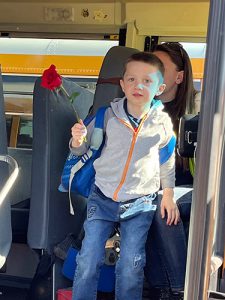 An elementary age boy wearing blue jeans and a gray hoodie holds a red rose as he gets off a bus.