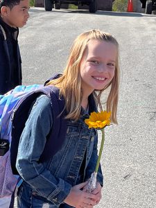 An older elementary school girl withlong blonde hair, wearing a denim jacket and a backpack, smiles as she holds a yellow sunflower.