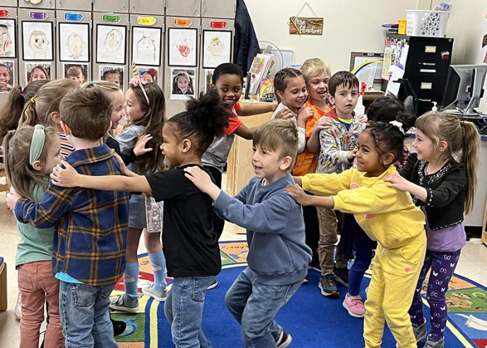A group of about 15 pre-K students go around in a circle, with hands on the shoulders of the person in front of them.