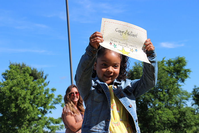 A little girl wearing a yellow print dress and a blue denim jacket smiles broadly. She is holding up a certificate that says Congratulations! And has multi-colored stars on it. There is a woman in the background smiling and clapping.