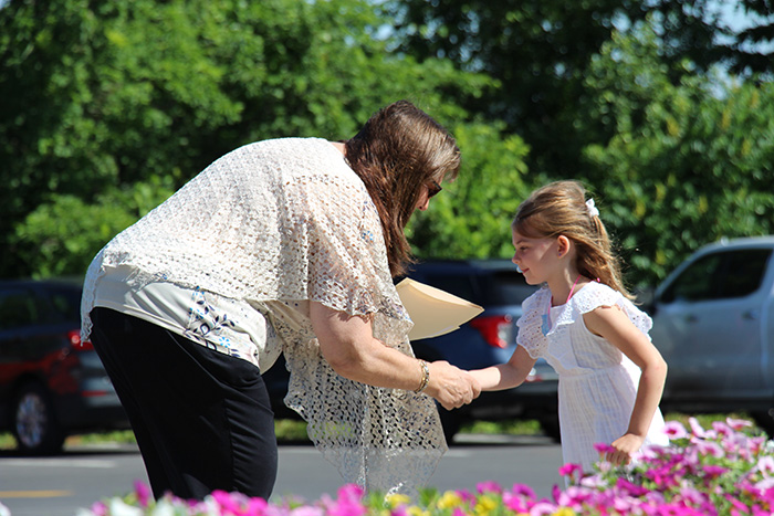 A girl with long brown hair, wearing a white dress, shakes hands with a woman with brown hair, wearing a beige shirt and black pants.