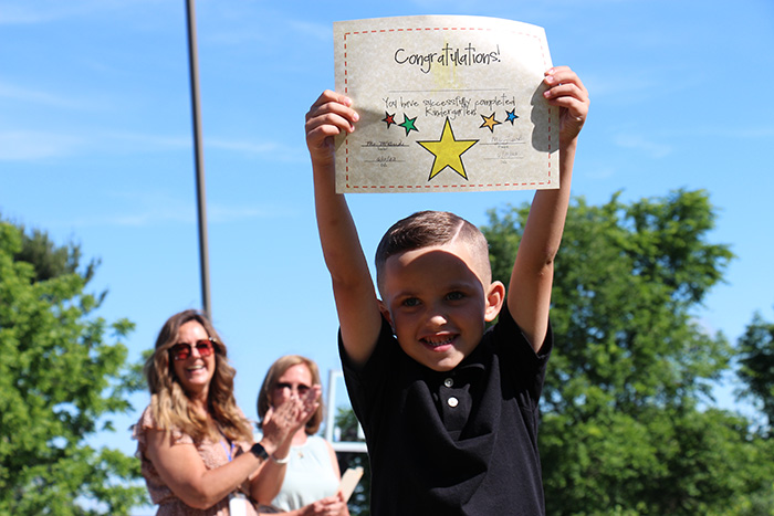 A little boy wearing a black polo shirt with short brown hair smiling broadly, holds up a certificate that says Congratulations! That also has several multi-colored stars. There are two women in the background smiling and clapping. There are also trees in the background and a bright blue sky.
