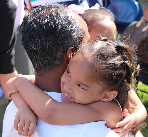 A little girl with long dark hair that is pulled back smiles and hugs a man with short salt and pepper hair.