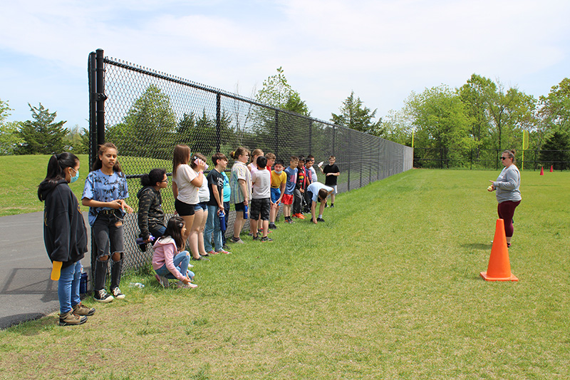 A group of elementary school students stand against a fence and listen to their teacher who is on the right.