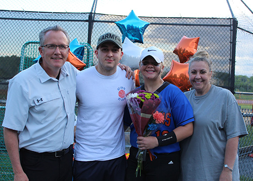 Blue and orange stars balloons behind four people. A man, a young man, a high school senior girl wearing a softball uniform and holding flowers, and a woman on the end. All are smiling.