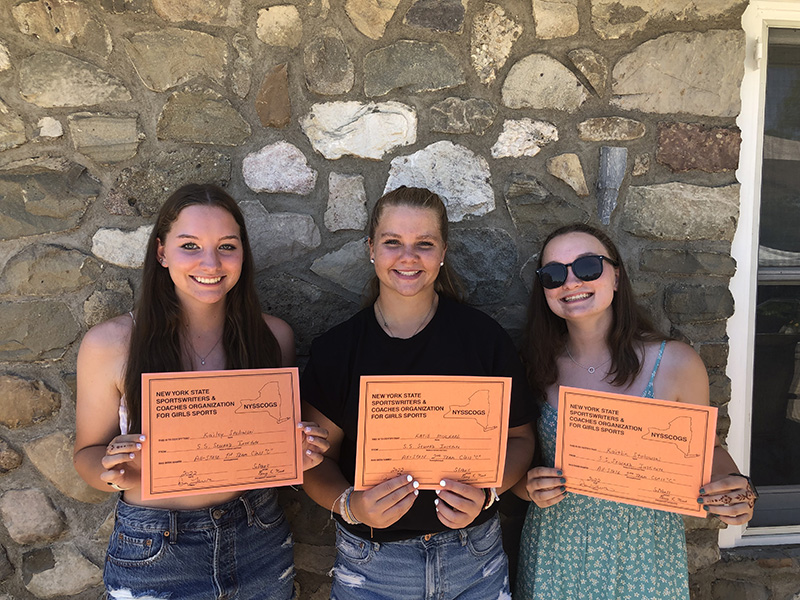 Three young women stand in front of a stone wall, each holding an orange certificate. Each has long hair and is smiling.