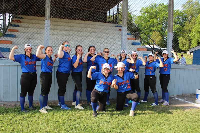 Twelve young women all dressed in blue and black softball uniforms, flex their muscles in front of stands at a baseball field. They are smiling.