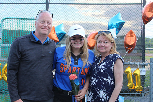 Three people in front of blue and orange star balloons. A man n the right, wearing a dark shirt, a high school senior in the center wearing a softball uniform - blue with orange letters that says Spartans - and a woman wearing a blue and white blouse on the right.