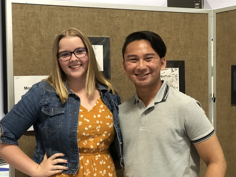 A young woman in a yellow and white dress and a denim jacket, with longer blonde hair and glasses smiles. She is on the left. On the right is a young man with short dark hair, wearing a light colored polo shirt. He is smiling too.