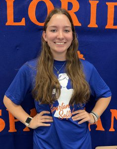A high school girl with long brown hair stands in front of a banner that says Florida Spartans. She is smiling, has her hands on her hips, and is wearing a blue tshirt that says Spartan Softball.