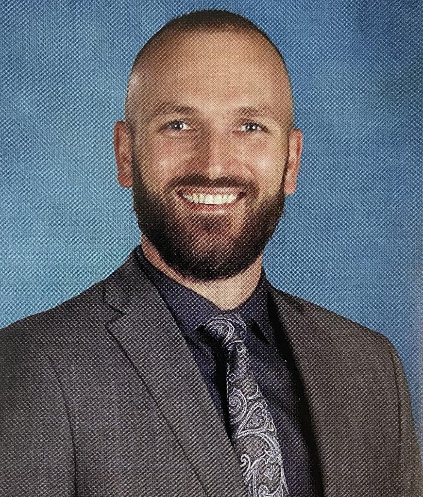 A man with a beard and mustache, smiles. He is wearing a gray jacket, blue button down shirt with a printed tie.