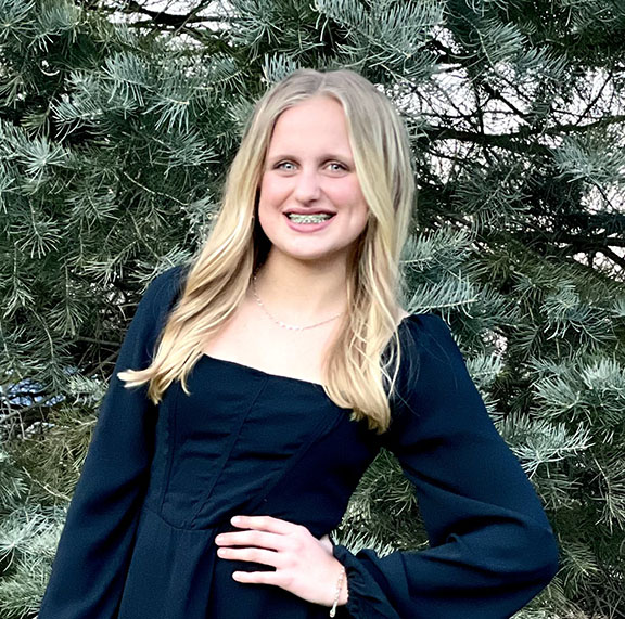 A girl with blond hair wearing a dark blue dress poses in front of a pine tree