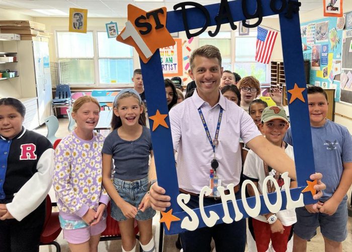 A teacher holds a First Day of School frame with his class around him