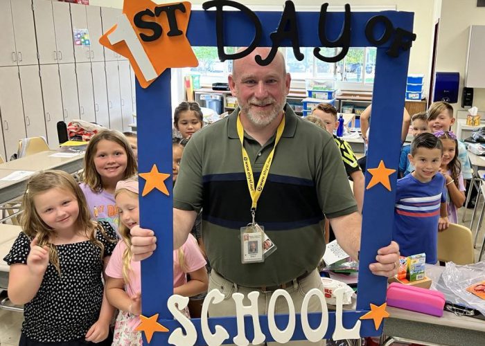 A teacher holds a first day of school frame as her class stands behind him.