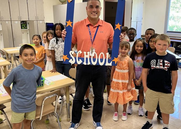 A teacher holds a First Day of School frame with his class around him