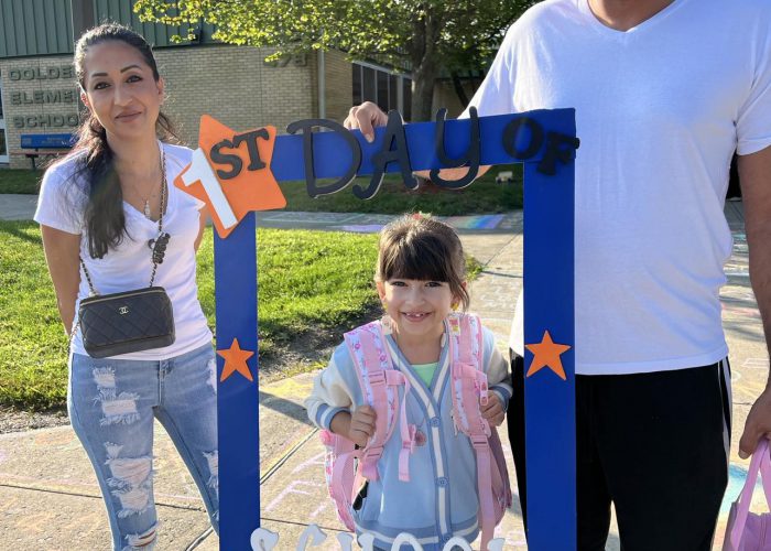 A girl stands inside a first day of school frame as two adults stand near