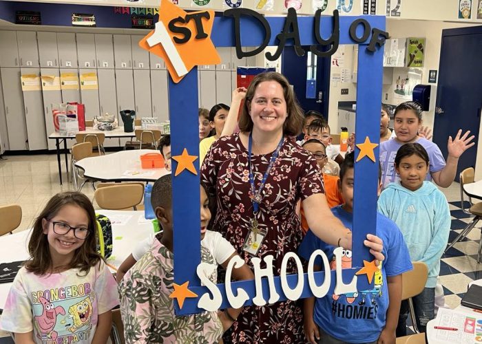 A teacher holds a First Day of School frame with her class around her