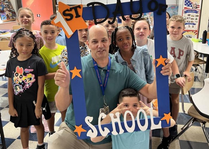 A teacher holds a First Day of School frame with his class around him