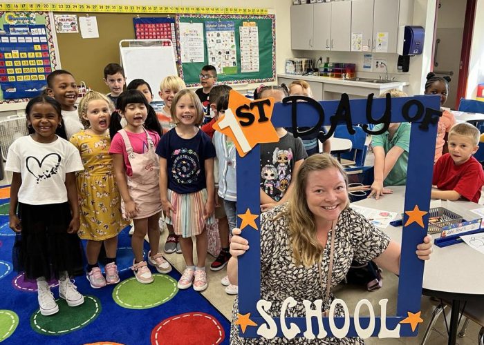 A teacher holds a first day of school frame as her class stands behind her.