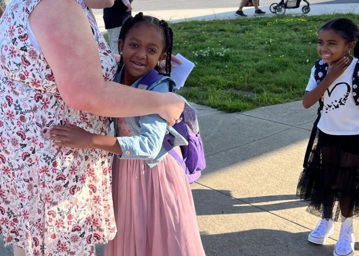 A teacher hugs a student as another student stands near