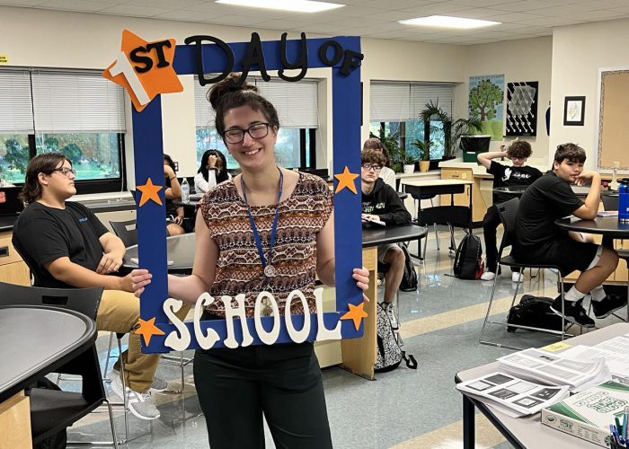 A person poses with a first day of school frame as student sit behind her in a class room.