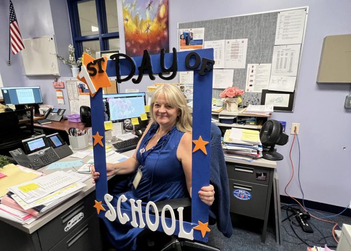 A person seated at a desk poses with a first day of school frame