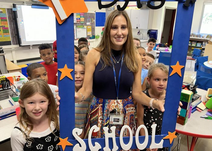 A teacher holds a First Day of School frame with her class around her