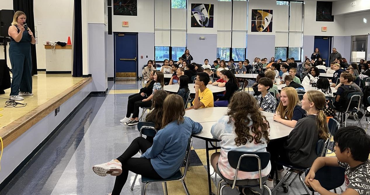 A person stands on a stage a students watch while seated at round tables